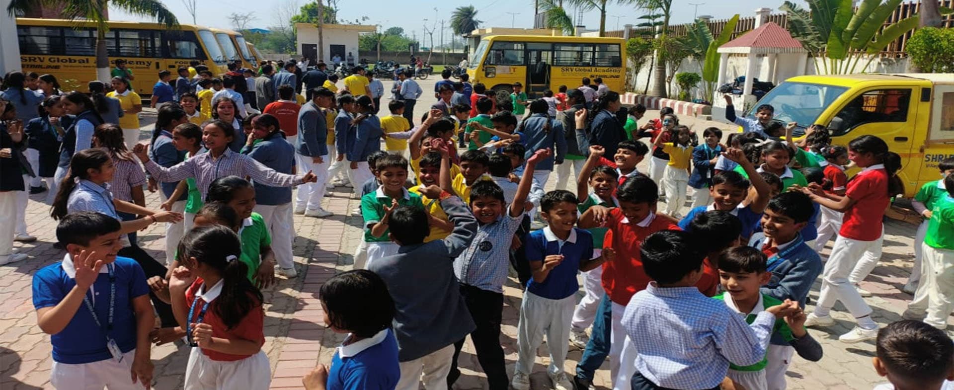 Students in house colours celebrating Holi in the school courtyard, buses behind them